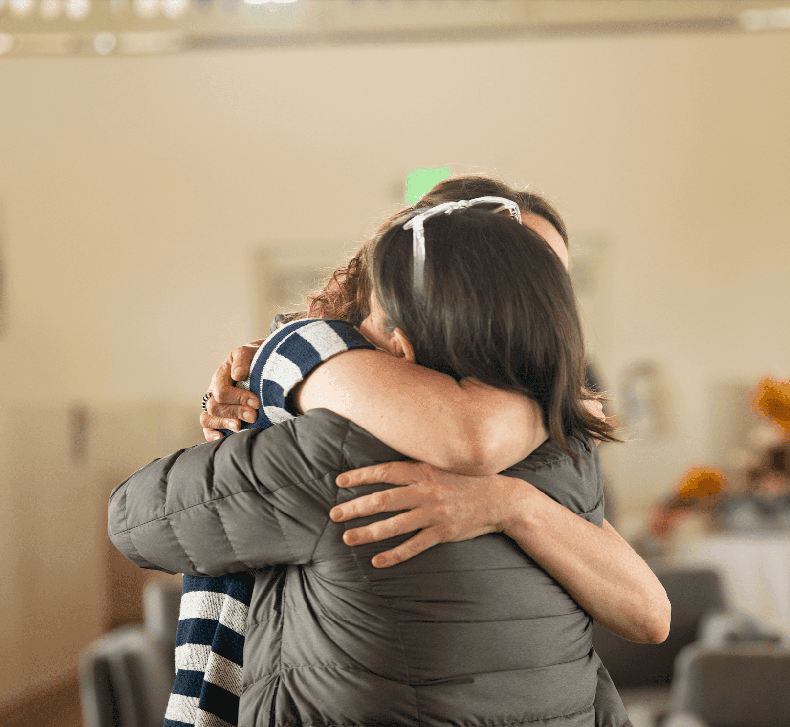 Image of two people sharing a hug, their faces not visible. One is brown haired with a green puffer jacket, the other wears a short sleeved striped shirt.